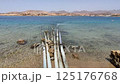 Multiple metal pipes extend from a sandy shore into transparent turquoise sea, surrounded by coral rocks and with distant mountains on the horizon 125176768