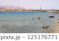 Scenic seashore with clear shallow waters, rocky shoreline, and a resort town with mosque minarets and mountains in the background under a hazy sky 125176771