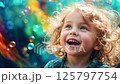 Joyful laughing toddler girl with curly blonde hair and bright blue eyes against colorful bokeh background with bubbles, expressing pure childhood happiness and wonder 125797754