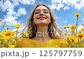 Young woman with glasses smiling with closed eyes in yellow dress among blooming sunflowers on sunny summer day, expressing joy and freedom against blue sky with white clouds 125797759