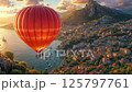 Hot air balloon flying over coastal mediterranean town with mountain backdrop at golden sunset, aerial view of harbor filled with boats and hillside with traditional architecture 125797761