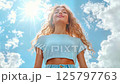 Young woman with blonde curly hair wearing blue crop top smiling with closed eyes against sunny sky and white clouds, expressing joy and freedom in summer weather 125797763