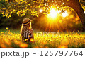 Young woman in straw hat sitting among wild yellow flowers in golden sunlit meadow at sunset, peaceful nature scene with sunlight streaming through tree branches 125797764