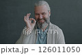 Slow motion portrait of glad mature man showing OK hand gesture smiling looking at camera on gray color background. People and emotion concept. 126011050