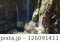 Rocky landscape with verdant moss, cascading waterfall streaming down lush terrain under bright sunlight, creating tranquil wilderness scenery 126091411