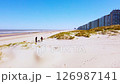A scenic beach landscape featuring pristine sand dunes, calm waters, and adjacent modern residential buildings against a blue sky with people walking on the beach 126987141
