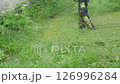 A worker is mowing overgrown grass and weeds along a building with a string trimmer, tidying up a yard by cutting the overgrowth. 126996284
