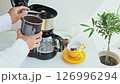 A person uses a spoon to add ground coffee to a drip coffee maker, to prepare a beverage next to a coffee cup and an olive tree. 126996294