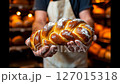 Freshly baked braided bread held by a baker in apron, with flour on hands. Warm artisan bakery setting, concept of homemade food, traditional craftsmanship, and authentic baking culture. 127015318