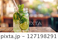 Fresh lime and mint drink in a mason jar with ice cubes on a rustic table. Summer refreshment concept. Vibrant colors, close-up shot in a blurred outdoor cafe background with natural lighting. 127198291