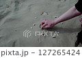 Close-up of hand drawing a heart shape on sandy beach surrounded by seashells in serene coastal environment 127265424