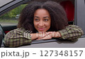 Portrait of attractive African American woman with stylish Afro hairstyle sitting in drivers seat, leaning on opened car window and looking with cheerful smile while enjoying road trip in city. 127348157