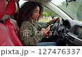 Positive charming black female driver with stylish haircut sitting in vehicle, recording voice message on cellphone while driving car in traffic jam in rush hour. 127476337