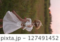 Young blonde girl in white dress is spinning cheerfully in summer field. 127491452
