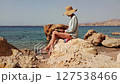 A young woman sits on a stone by the sea and reads a book. She has a straw sunhat.  127538466