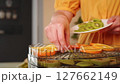 Woman carefully placing kiwi slices on a dehydrator tray already filled with orange slices, preparing healthy snacks 127662149