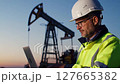 Profile of Male Engineer at Oil Well with Laptop Hardhat and Safety Vest Against a Clear Sky at Dusk 127665382