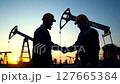 Silhouetted Oil Workers in Orange Hardhats Shaking Hands at Sunset with Rigs and Blue Skies and Fields 127665384