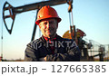 Smiling Oil Field Worker with Red Hardhat in Front of Oil Derrick against Blue Sky on Bright Sunny Day 127665385