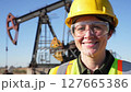 Smiling Woman Oil Field Worker in Yellow Hard Hat Clear Safety Glasses and High Visibility Vest with Oil Pump and Clear Blue Sky 127665386