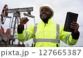 Successful African American Engineer with Tablet at Oil Field with Pump Jack in a White Hardhat and a Yellow Safety Jacket 127665387