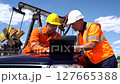Two Technicians in Hard Hats Collaborating on Laptop Near Oil Pump Under Blue Sky 127665388
