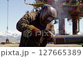 Welder Working on Steel Pipe Wearing a Helmet Against Offshore Platform And Mountain Backdrop Under a Bright Blue Sky 127665389