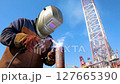 Welder Working with Metal on Construction Site Under Clear Blue Sky, Wearing Protective Gear and Welding Equipment with Sparks 127665390
