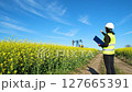 Woman Inspecting Oil Derrick in Yellow Canola Field Under a Clear Blue Sky Holding Blue Clipboard and Wearing Hard Hat 127665391
