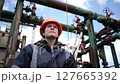 Woman in Blue Jacket and Orange Hardhat Holds Walkie Talkie by Weathered Industrial Pipes Under Partly Cloudy Sky 127665392