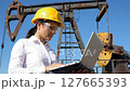 Woman In Safety Helmet Operates Laptop Near Rust Colored Oil Rig With Clear Blue Sky Industrial Equipment And Protective Eyewear 127665393