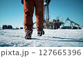 Worker in Orange Uniform Walks on Snowy Ground Near Oil Production Site Under Bright Blue Sky On Cold Winter Day 127665396