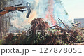 Large yellow excavator operating near burning pile of tree roots, branches, and forest debris with colorful smoke against background of trees and white building. Mechanical site preparation and waste 127878051