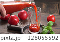 Hands pouring homemade tomato juice from glass bottle into mug  fresh tomatoes and basil on table. 128055592
