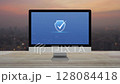 Security shield with check mark icon on desktop computer monitor screen on wooden table over blur of cityscape on warm light sundown, Technology internet cyber security and anti virus online concept 128084418