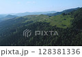 Aerial view of a dirt road winding through lush green hills and dense forest, with distant mountain ranges under a clear blue sky on a summer day. 128381336