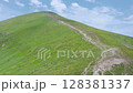 Aerial view of a green mountain slope with a winding trail leading to the summit under a blue sky with scattered clouds on a clear summer day. 128381337