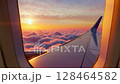 Airplane Window View Above the Clouds at Sunset. Beautiful purple and orange sky. Aerial view of Cloudscape. Airplane wing in sky. View through porthole from passenger seat 128464582