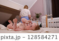 A caregiver gently changes a toddler's diaper indoors, ensuring the child is comfortable on a soft bed. Natural light fills the room, highlighting the intimate moment of care. 128584271