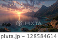 A breathtaking sunrise illuminates a tranquil coastline, with soft waves reflecting the golden light. Rugged mountains frame the horizon under a sky painted in pastel hues and drifting clouds. 128584614