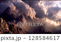 A breathtaking view of jagged mountain peaks shrouded in dramatic, swirling mist and clouds, with rays of sunlight breaking through, creating an ethereal and awe-inspiring landscape scene. 128584617