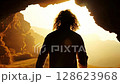 View from behind of a man with long, slightly curly hair.He is exiting a dark cave, and his silhouette is surrounded by bright light. The atmosphere has cinematic lighting and warm tones 128623968