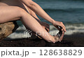 Closeup of woman bare feet, female shakes feet off black sand on beach. Barefoot woman with long legs sitting on beach of Pacific Ocean on background of waves. Slow motion shot, natural light 128638829
