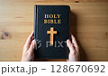 Close up of a person's hands holding the Holy Bible with a wooden cross on a table. Concept of Christian faith, spirituality, and worship. 128670692