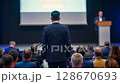 Speaker on stage giving a presentation to an audience at a business conference. People at a corporate event listening to a lecture in a dark hall with a large screen.. 128670693