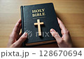 Close up of a person's hands holding the Holy Bible with a wooden cross on a table. Concept of Christian faith, spirituality, and worship. 128670694