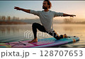 Man practicing warrior yoga pose on paddleboard at dawn in misty calm lake surrounded by trees 128707653