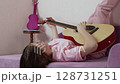 Closeup of young guitarist playing acoustic guitar lying on back on sofa with legs up against wall. Musician is experimenting with guitar in home. Woman is wearing jeans and polo shirt. Handheld shot 128731251