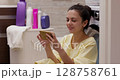 brunette woman using smartphone while laundry is in washing machine, woman sitting on the floor near washing machine in bathroom 128758761