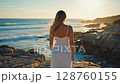 Woman in white dress enjoys tranquil ocean sunset on rocky coastline horizon 128760155